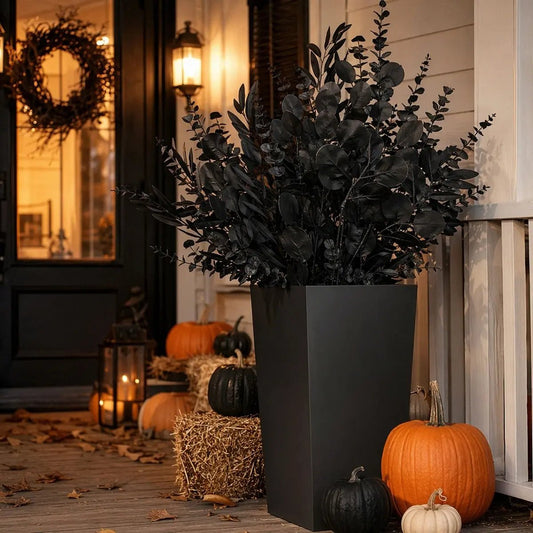 Mixed black faux eucalyptus stems in a tall black planter on a front porch Halloween display with orange and black pumpkins, hay bales, candle lanterns, and fall leaves at dusk