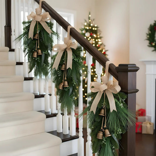 Three Christmas pine needle teardrop swags with burlap bows and hanging vintage brass bells, hung at intervals along a dark wood staircase railing, with a fully decorated Christmas tree glowing in the background of a classic white and wood holiday home interior