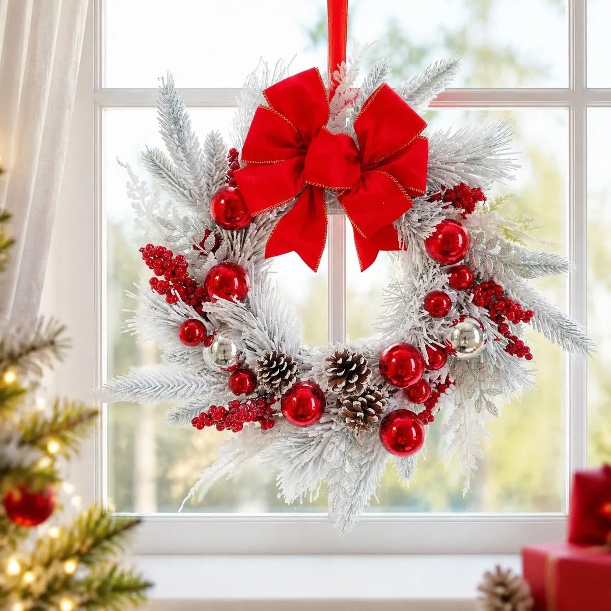 17.7 inch snow flocked Christmas wreath with red velvet gold-trim bow, shiny red and silver ball ornaments, abundant red berry clusters and snow-dusted pinecones on white flocked pine branches, hung by a red ribbon in the center of a large white-framed window with sheer white curtains, with a lit Christmas tree and red gift boxes visible to the left in a bright white Christmas living room