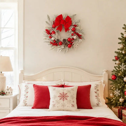 17.7 inch snow flocked Christmas wreath with red velvet gold-trim bow, shiny red and silver ball ornaments, red berry clusters and snow-dusted pinecones, hung on a warm greige bedroom wall above a white wood bed headboard dressed with white linen bedding, red velvet accent pillows and white snowflake embroidered pillow shams, with a white ceramic house lamp on the nightstand and a fully decorated Christmas tree with red and silver ornaments beside the bed