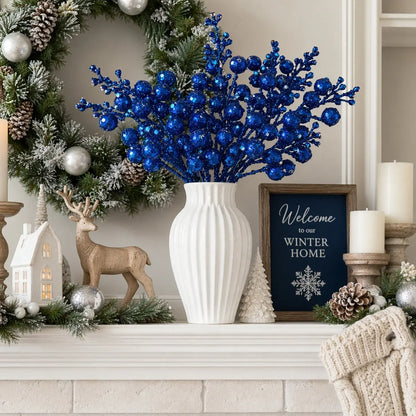 Glitter blue artificial berry stems in a white ribbed vase as a fireplace mantel centerpiece, surrounded by a frosted pine and silver ornament wreath, wooden reindeer figurine, white ceramic village house, pine cones and a Welcome to our Winter Home chalkboard sign