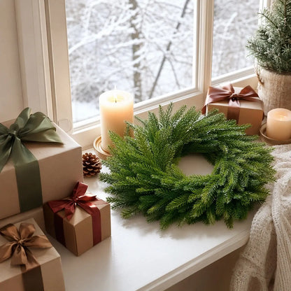 Artificial Norfolk pine candle ring wreath displayed on a window sill with a pillar candle, pine cone and wrapped Christmas gifts, with snowy winter trees visible through the window
