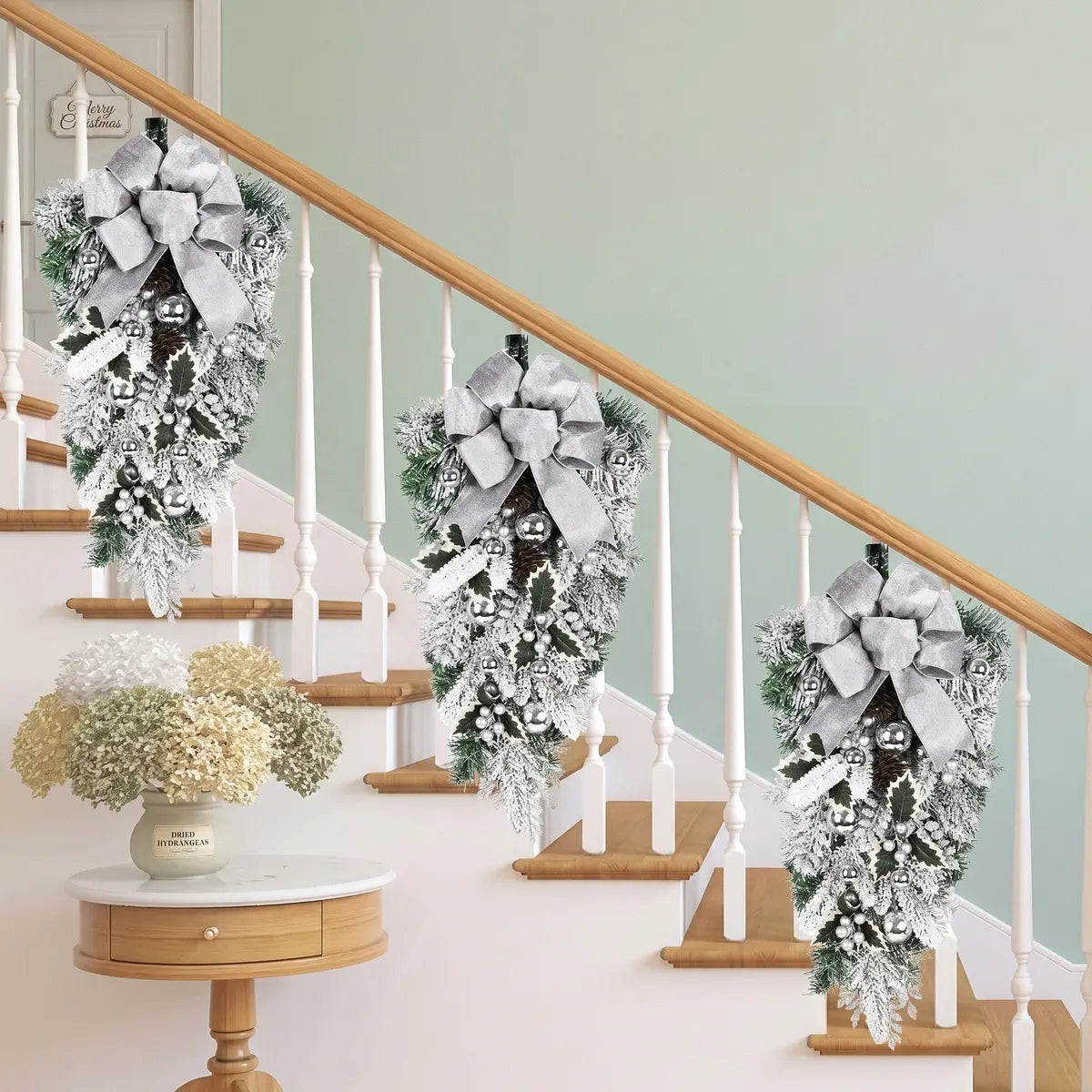 Three white Christmas teardrop swags with silver bows, silver ball ornaments and frosted holly leaves, hung on a white staircase railing banister in a bright farmhouse hallway with a Merry Christmas sign and dried hydrangea arrangement
