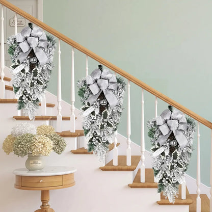 Three white Christmas teardrop swags with silver bows, silver ball ornaments and frosted holly leaves, hung on a white staircase railing banister in a bright farmhouse hallway with a Merry Christmas sign and dried hydrangea arrangement
