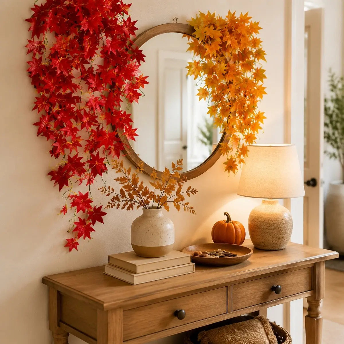 Red and yellow artificial maple leaf garlands draped around round mirror on entryway console table with pumpkin, fall home decor