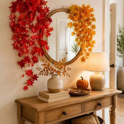 Red and yellow artificial maple leaf garlands draped around round mirror on entryway console table with pumpkin, fall home decor