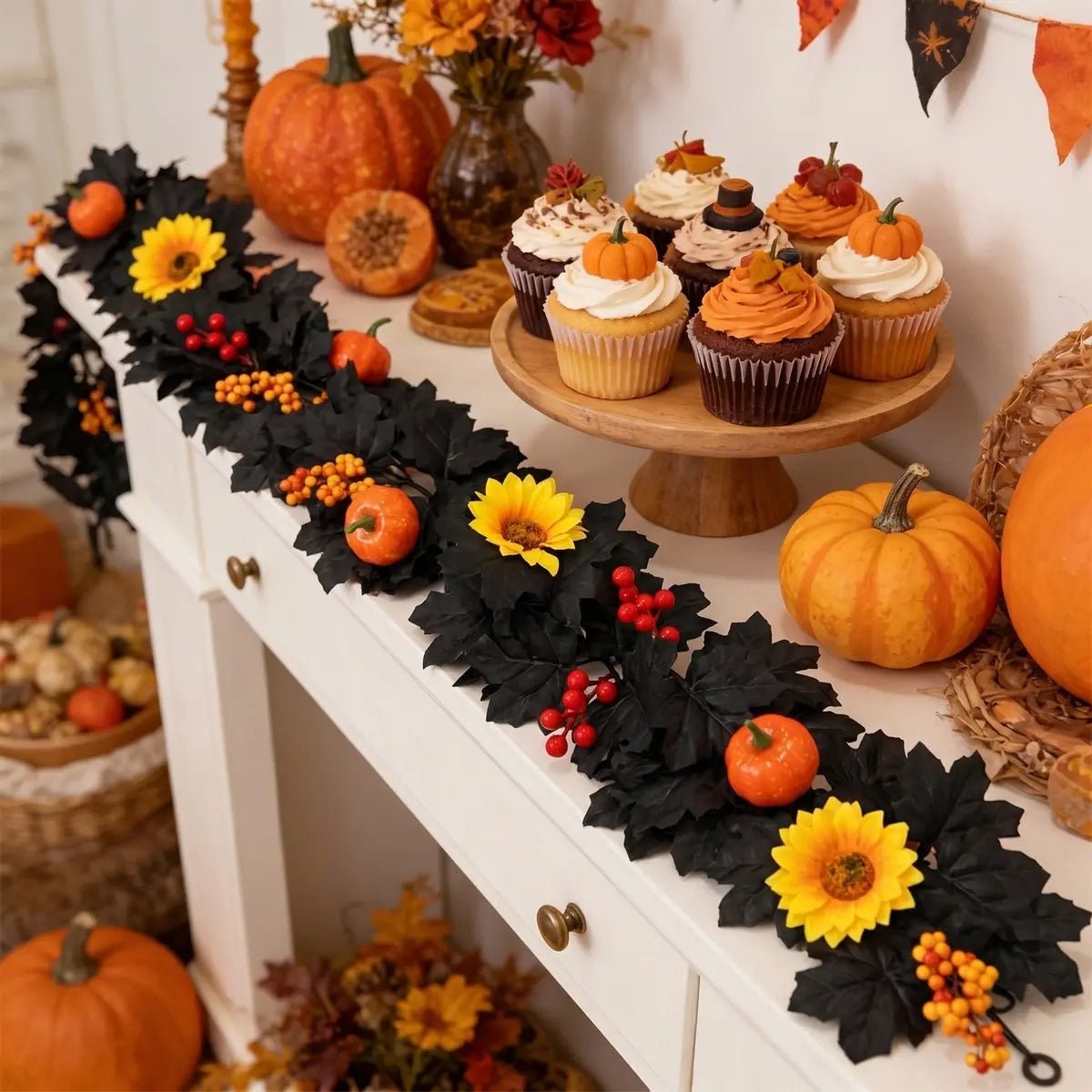 Black autumn garland with yellow sunflowers, orange pumpkins, and red berry clusters draped along a white console table with Halloween cupcakes, orange pumpkins, and wicker baskets for fall harvest party display