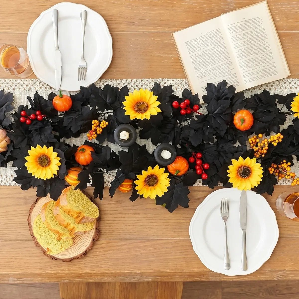 Black autumn garland with yellow sunflowers, orange pumpkins, and red berries used as a table runner on a wooden dining table with white plates, candles, and bread board for Thanksgiving harvest table decor