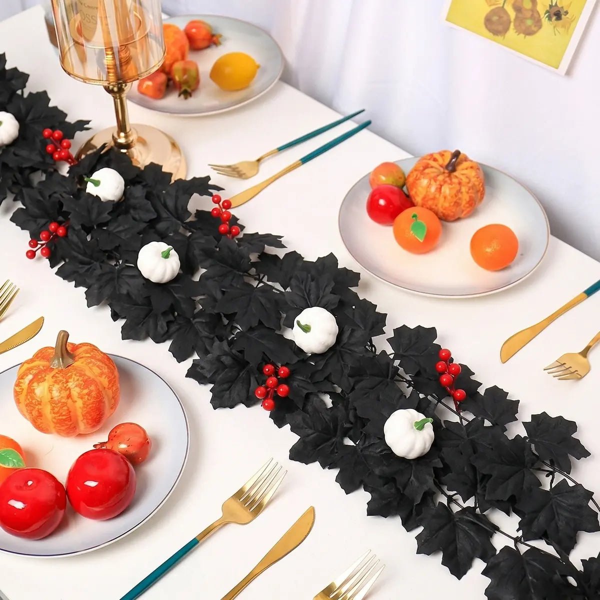 Black maple leaf garland with white pumpkins and red berry accents used as a table runner on a white dining table with gold cutlery, orange pumpkins, and fruit for Thanksgiving harvest decor
