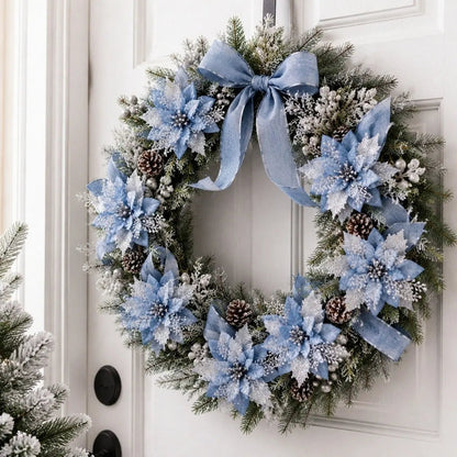 Light blue glitter artificial poinsettia picks clipped onto a frosted pine Christmas wreath with natural pinecones, silver berry clusters and a large dusty blue linen bow, hung on a white six-panel front door with a black iron hook, beside a snow-dusted potted Christmas tree