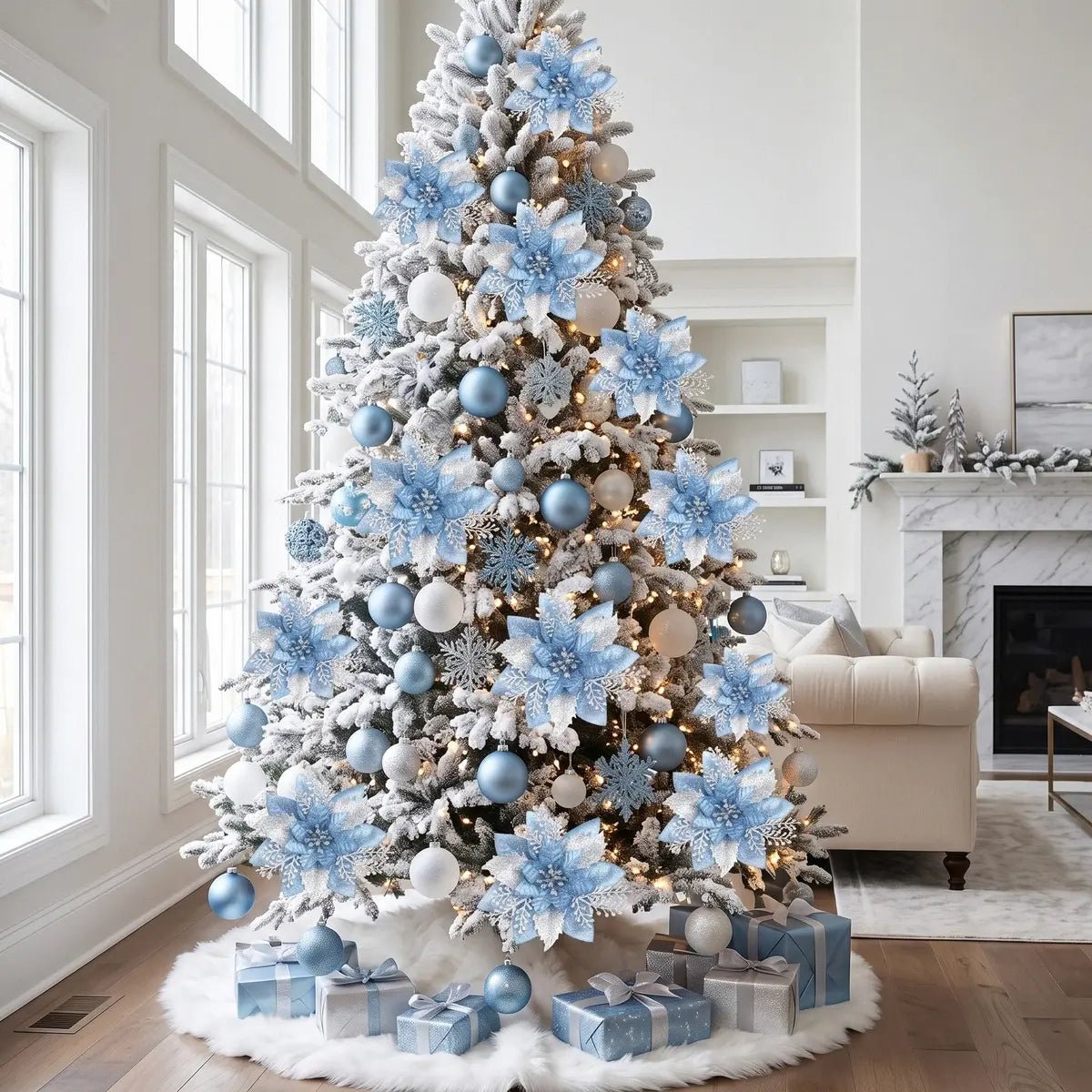 Tall flocked white Christmas tree fully decorated with light blue glitter artificial poinsettia picks, matte blue and white ball ornaments, white glitter snowflake ornaments and warm fairy lights, in a bright modern white living room with floor-to-ceiling windows, a white fur tree skirt and silver-wrapped Christmas gifts underneath, with a marble fireplace and frosted mini Christmas tree on the mantel in the background
