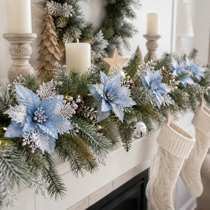 Light blue glitter artificial poinsettia picks clipped onto a frosted pine mantel garland with silver berry clusters, natural pinecones and silver jingle bells, draped across a white fireplace mantel with distressed wood candlestick holders, white pillar candles, a wooden star and a wooden Christmas tree, with cream knit Christmas stockings hanging below