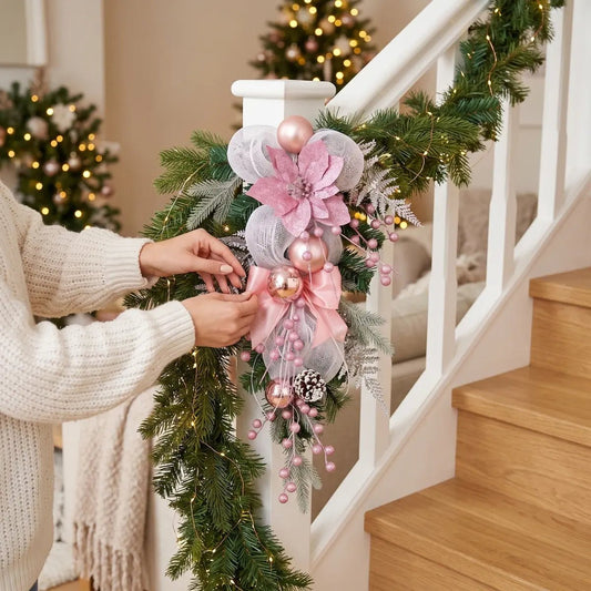 Pink Christmas teardrop swag with poinsettia, mesh bow and rose gold ball ornaments, displayed on white stair railing newel post with garland and lit Christmas tree in background
