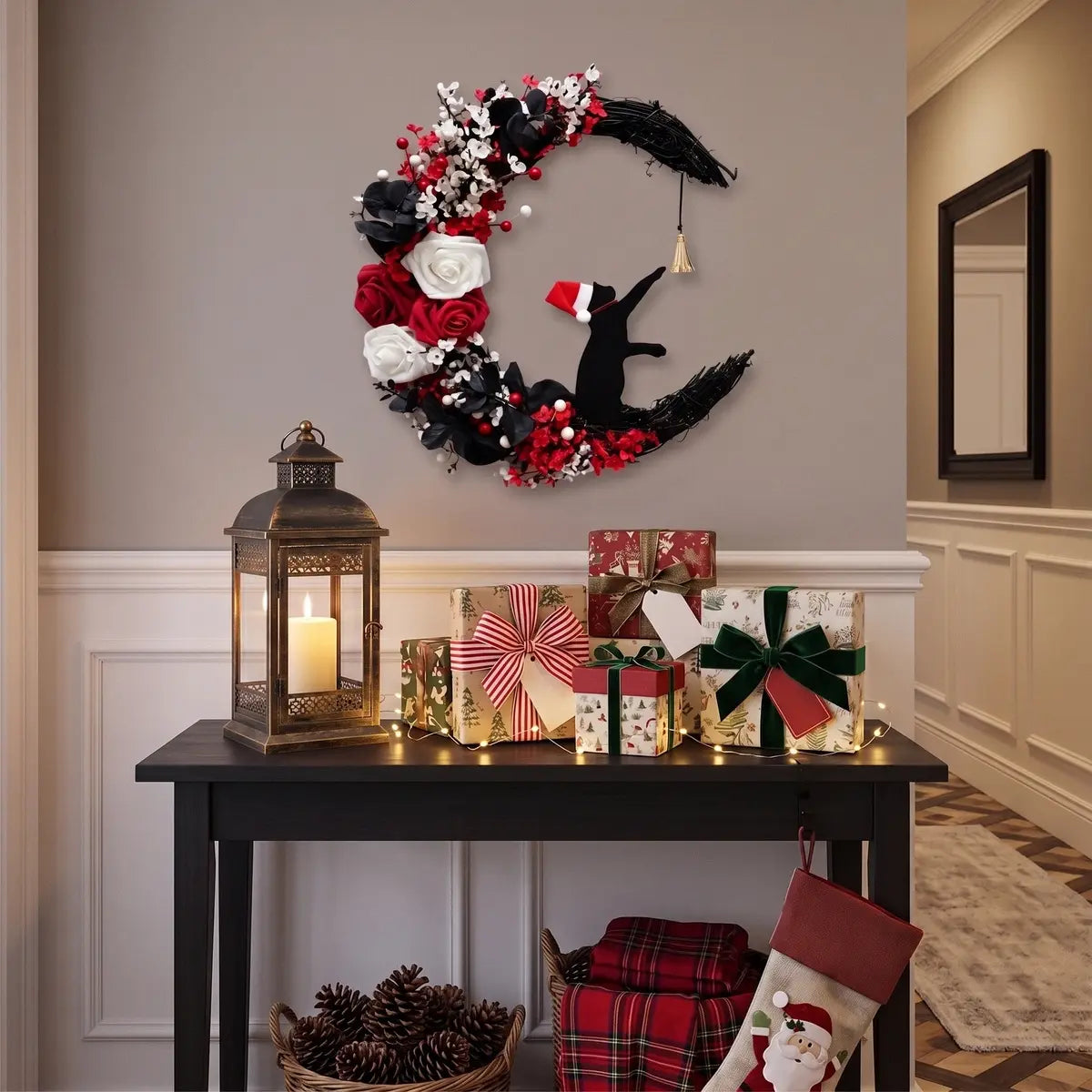 Christmas crescent moon black cat wreath with Santa hat, white and red roses and gold bell, displayed above a dark entryway console table with wrapped Christmas gifts, brass lantern and holiday stockings
