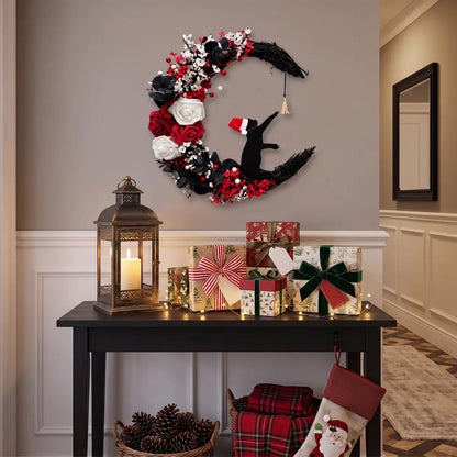 Christmas crescent moon black cat wreath with Santa hat, white and red roses and gold bell, displayed above a dark entryway console table with wrapped Christmas gifts, brass lantern and holiday stockings
