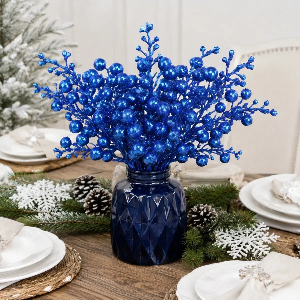 Glitter blue artificial berry picks bundled in a navy blue faceted glass vase as a Christmas dining table centerpiece, surrounded by frosted pine branches, white pine cones and snowflake decorations on a rustic wood farmhouse table with white place settings
