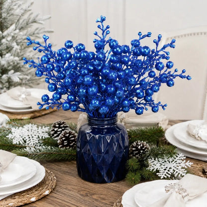 Glitter blue artificial berry picks bundled in a navy blue faceted glass vase as a Christmas dining table centerpiece, surrounded by frosted pine branches, white pine cones and snowflake decorations on a rustic wood farmhouse table with white place settings
