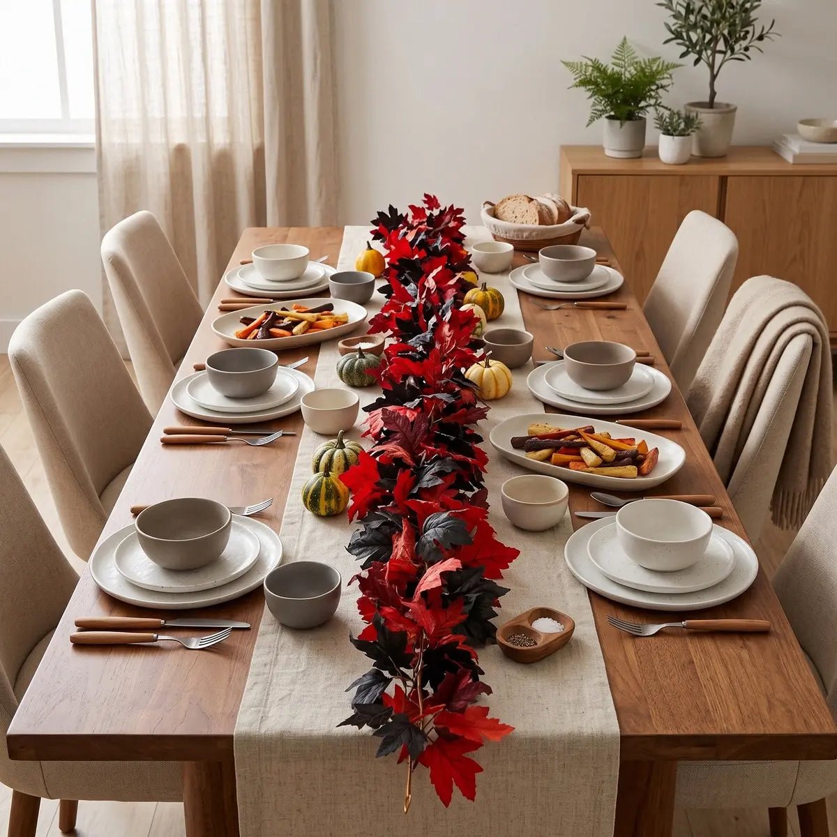Red and black maple leaf fall garland used as a table runner down the center of a wooden Thanksgiving dining table with white plates, mini green and orange pumpkins, bread basket, and linen runner