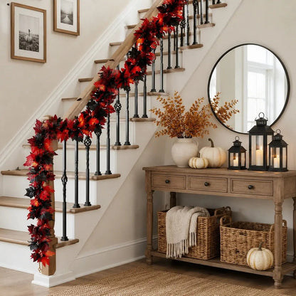 Red and black maple leaf fall garland wrapped along a black iron staircase banister with white pumpkins, black lanterns, and fall branches in a white vase on a wooden entryway console table for autumn home decor