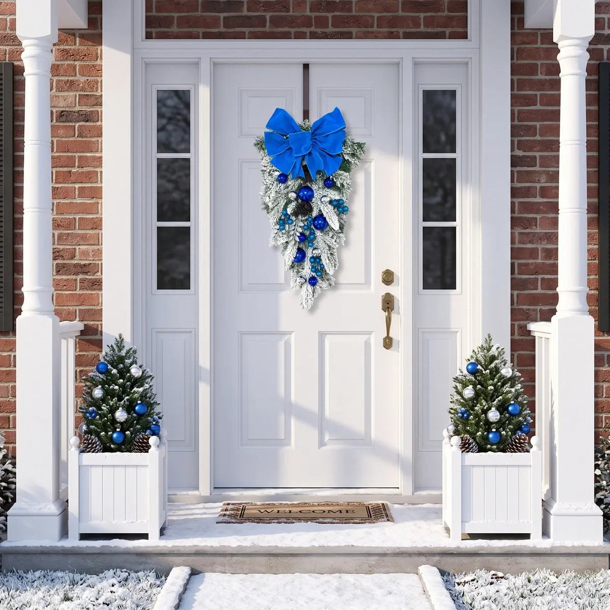 Blue Christmas teardrop swag with royal blue velvet bow, blue and silver ball ornaments and frosted pine branches, hung on a white front door with brick surround, flanked by white planter boxes with frosted mini Christmas trees decorated with blue and silver ornaments and pine cones, in a snowy winter setting
