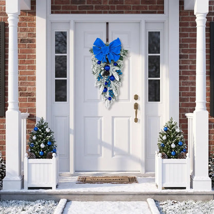 Blue Christmas teardrop swag with royal blue velvet bow, blue and silver ball ornaments and frosted pine branches, hung on a white front door with brick surround, flanked by white planter boxes with frosted mini Christmas trees decorated with blue and silver ornaments and pine cones, in a snowy winter setting
