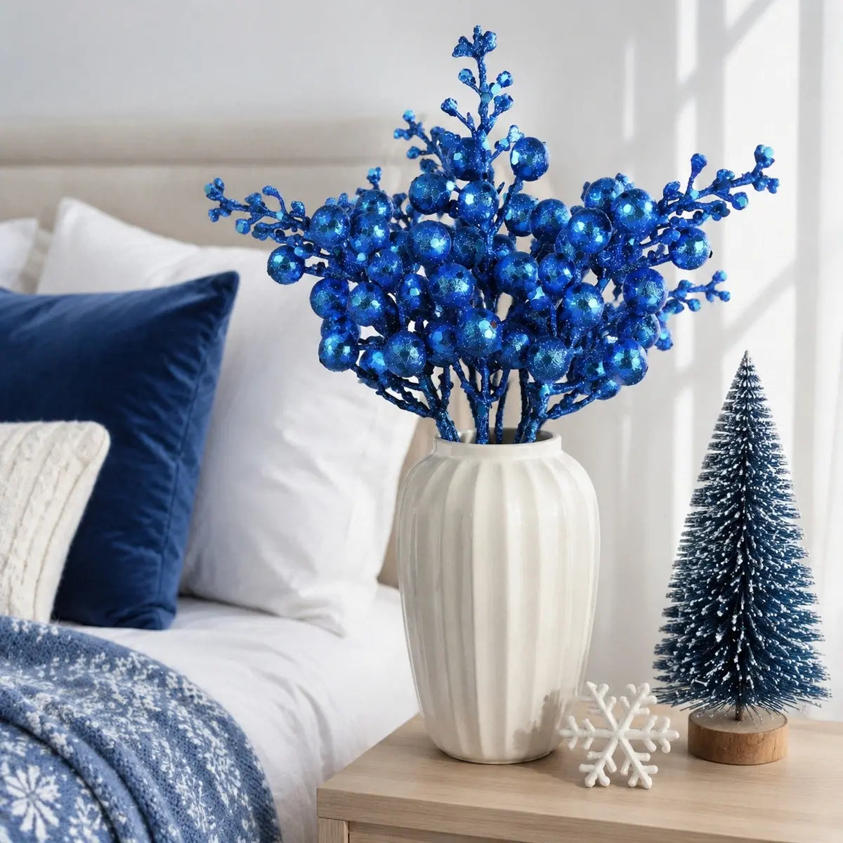 Glitter blue artificial berry stems in a white ribbed ceramic vase on a bedroom nightstand, styled beside a miniature frosted Christmas tree and white snowflake ornament, with navy blue velvet pillows and a blue snowflake print throw blanket in the background
