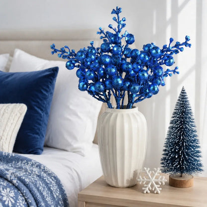 Glitter blue artificial berry stems in a white ribbed ceramic vase on a bedroom nightstand, styled beside a miniature frosted Christmas tree and white snowflake ornament, with navy blue velvet pillows and a blue snowflake print throw blanket in the background
