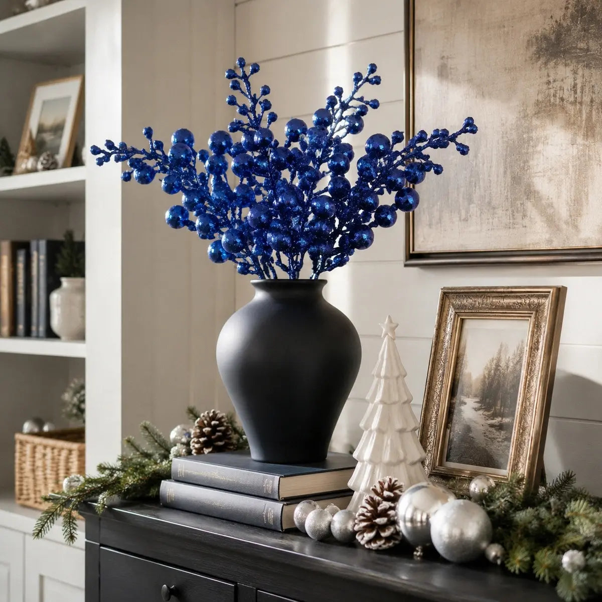 Glitter blue artificial berry stems in a matte black ceramic vase on a dark wood sideboard, styled with stacked books, pine cones, silver ball ornaments, a white ceramic Christmas tree and a framed winter landscape painting, with white shiplap wall and built-in bookshelves in background
