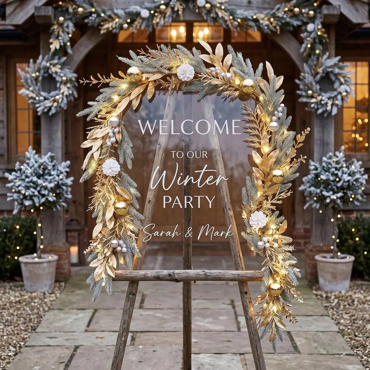 5.9FT Christmas gold leaves garland with gilded golden leaves, frosted pine branches, gold and silver ball ornaments, silver pearl berries and white pompom flowers with warm fairy lights, draped around a rustic wood arch frame with a Welcome to Our Winter Party acrylic sign, at an outdoor winter venue with frosted topiary trees and a stone manor house in the background