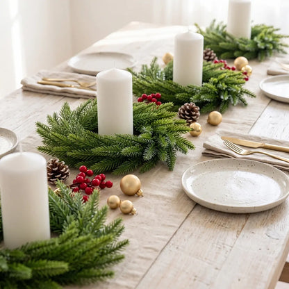 Artificial Norfolk pine candle ring wreath with white pillar candle, gold ball ornaments, red berries and pine cone, styled on a rustic white wood farmhouse dining table with gold cutlery
