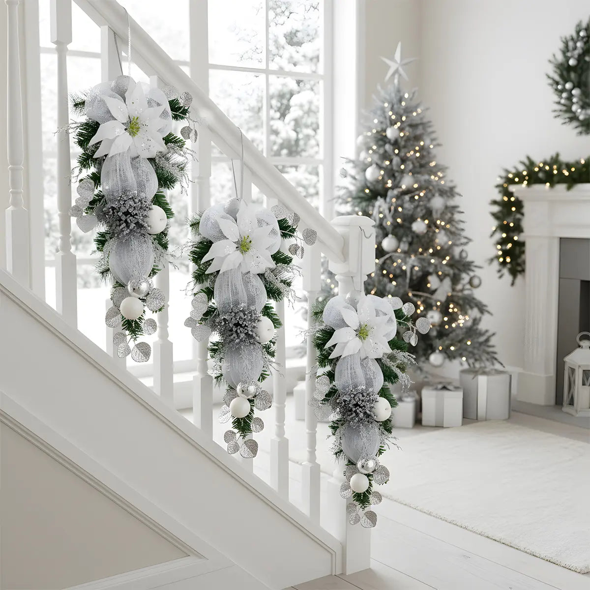 Silver white Christmas teardrop swag with glitter poinsettia and mesh bow, displayed on white stair railing with decorated Christmas tree in background
