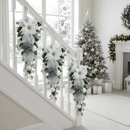 Silver white Christmas teardrop swag with glitter poinsettia and mesh bow, displayed on white stair railing with decorated Christmas tree in background

