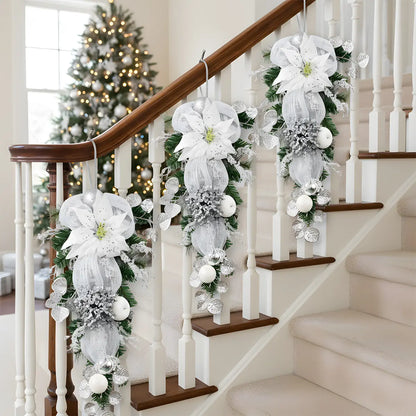 Three silver white Christmas teardrop swags with glitter poinsettia and mesh bows, decorating a wooden stair railing with lit Christmas tree in background
