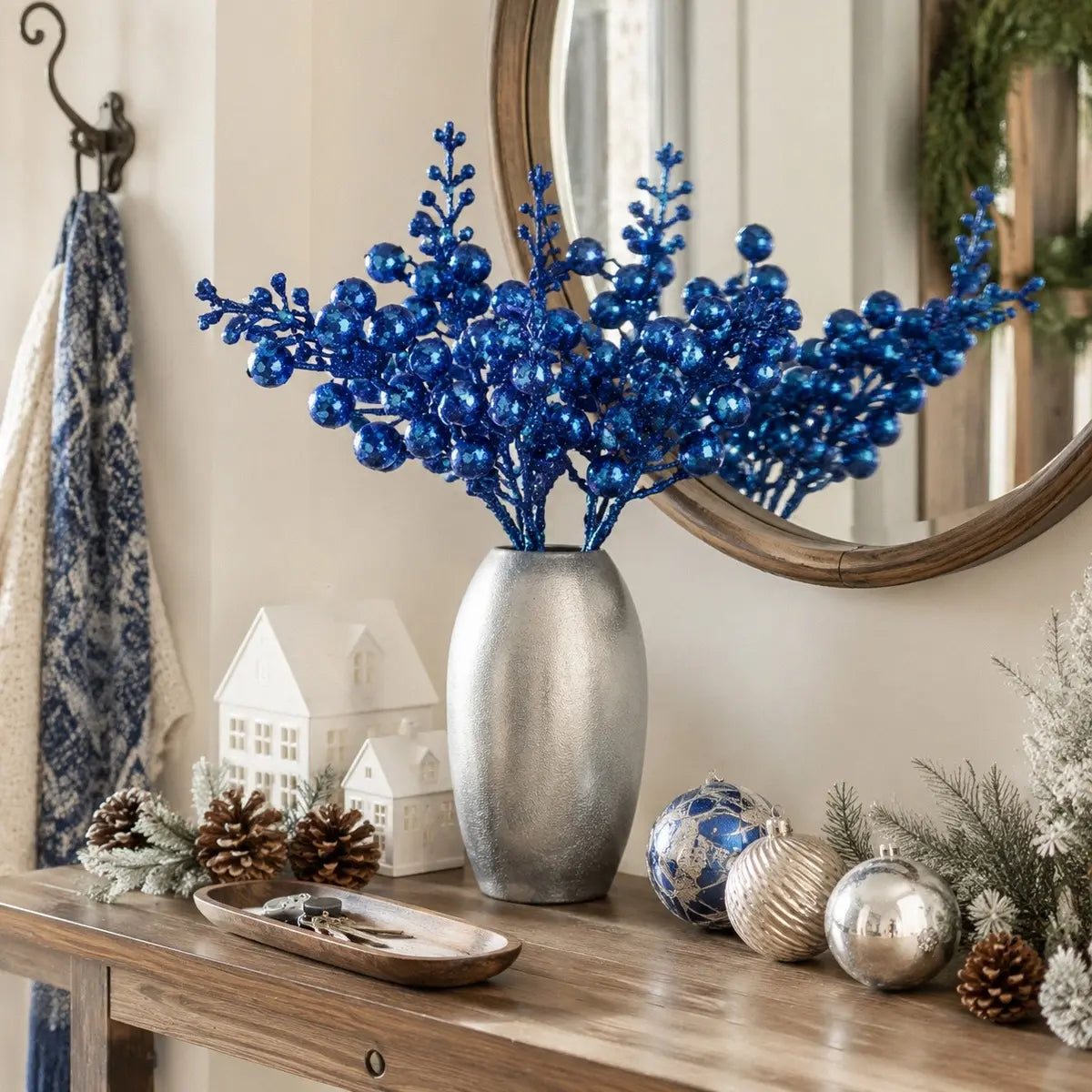 Glitter blue artificial berry stems displayed in a silver metallic vase on a rustic wood entryway console table, styled with pine cones, white ceramic village houses, blue and silver ball ornaments and a round mirror reflecting a Christmas wreath
