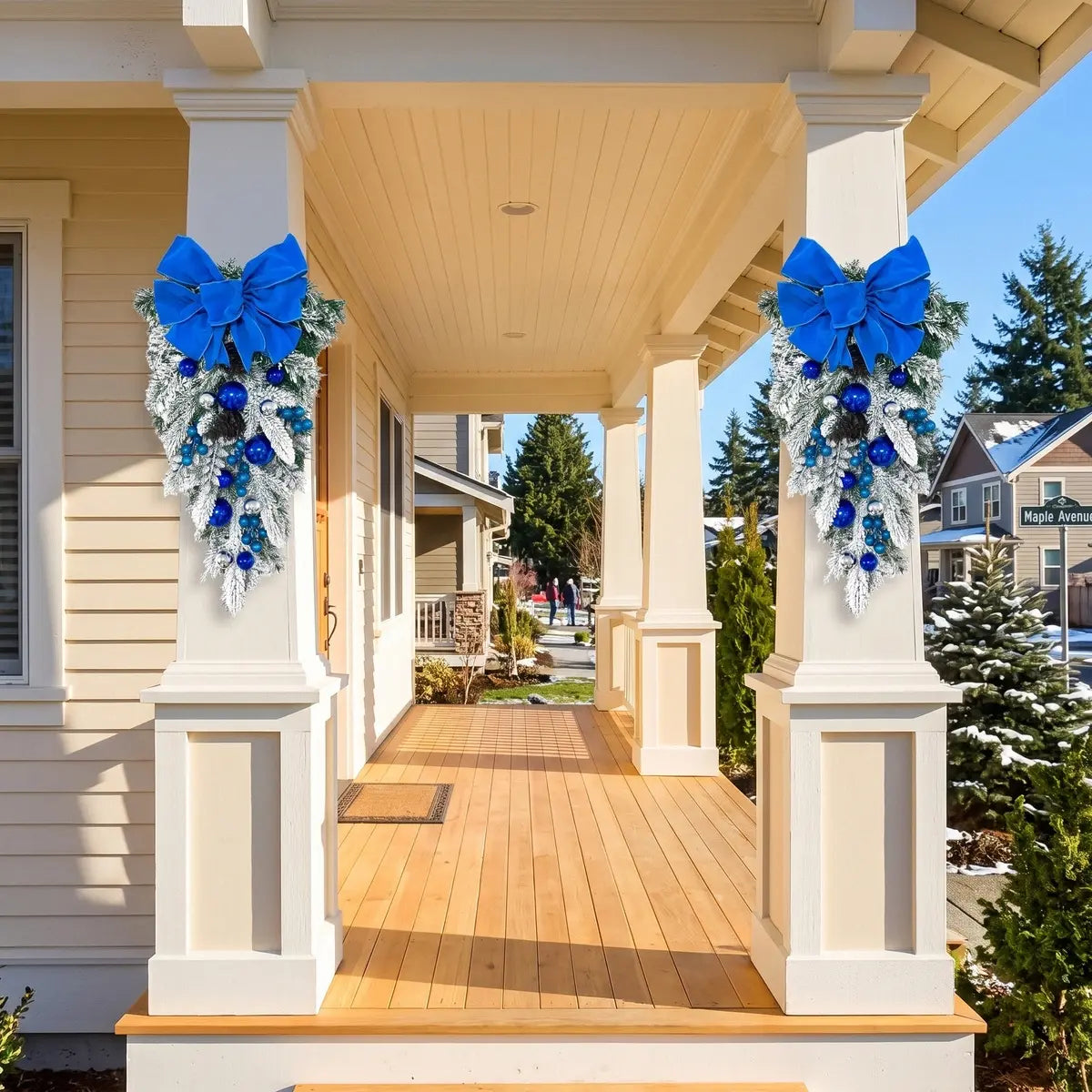 Pair of blue Christmas teardrop swags with royal blue velvet bows, blue and silver ball ornaments and frosted pine branches, hung on white porch columns of a suburban home in winter, with snow-covered yard and blue sky
