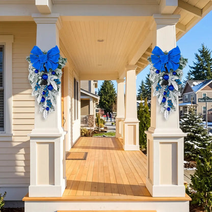 Pair of blue Christmas teardrop swags with royal blue velvet bows, blue and silver ball ornaments and frosted pine branches, hung on white porch columns of a suburban home in winter, with snow-covered yard and blue sky
