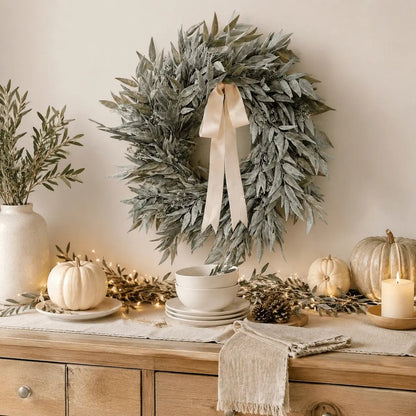 Artificial olive leaf wreath with cream ribbon bow hung on wall above rustic wood sideboard styled with white pumpkins, cream candles, fairy lights, and pinecones for fall harvest decor
