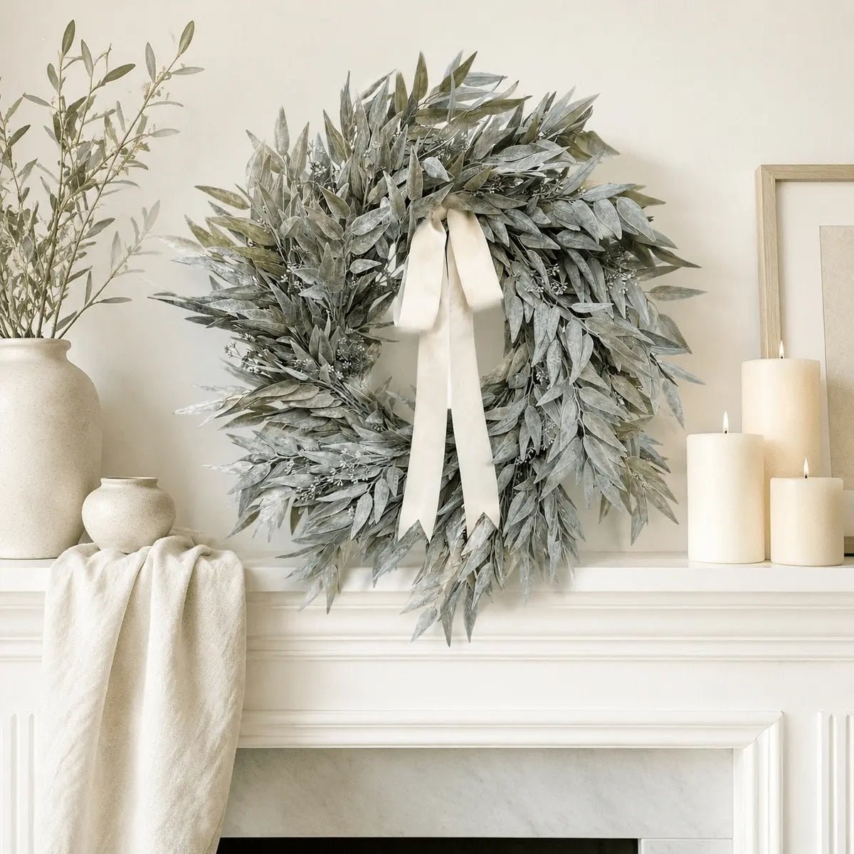 Artificial olive leaf wreath with cream satin bow displayed above white fireplace mantel with cream pillar candles and ceramic vases in neutral living room
