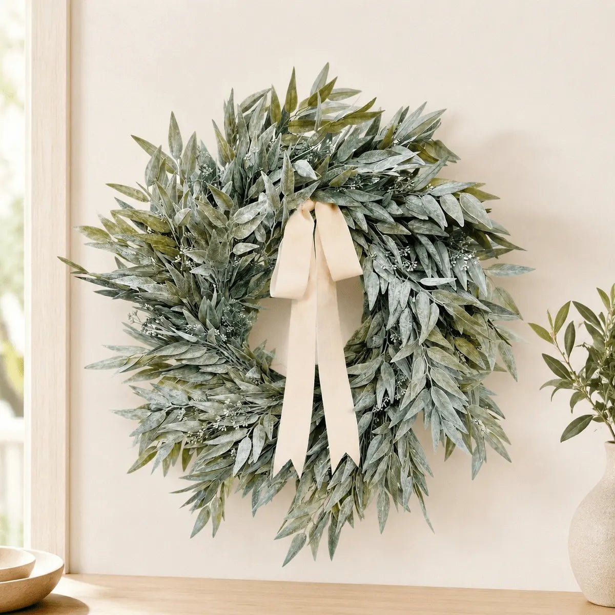 Artificial olive leaf wreath with cream satin ribbon bow hanging on white interior wall beside a window, styled with a small potted olive tree on a wooden shelf
