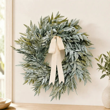 Artificial olive leaf wreath with cream satin ribbon bow hanging on white interior wall beside a window, styled with a small potted olive tree on a wooden shelf
