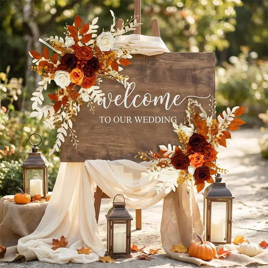 Burnt orange boho artificial flower swag decorating a rustic wedding welcome sign with lanterns and draped fabric
