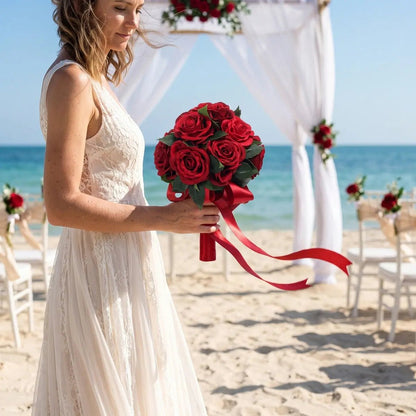 Bride holding artificial red rose bouquet with red satin bow at beach outdoor wedding ceremony, ocean backdrop
