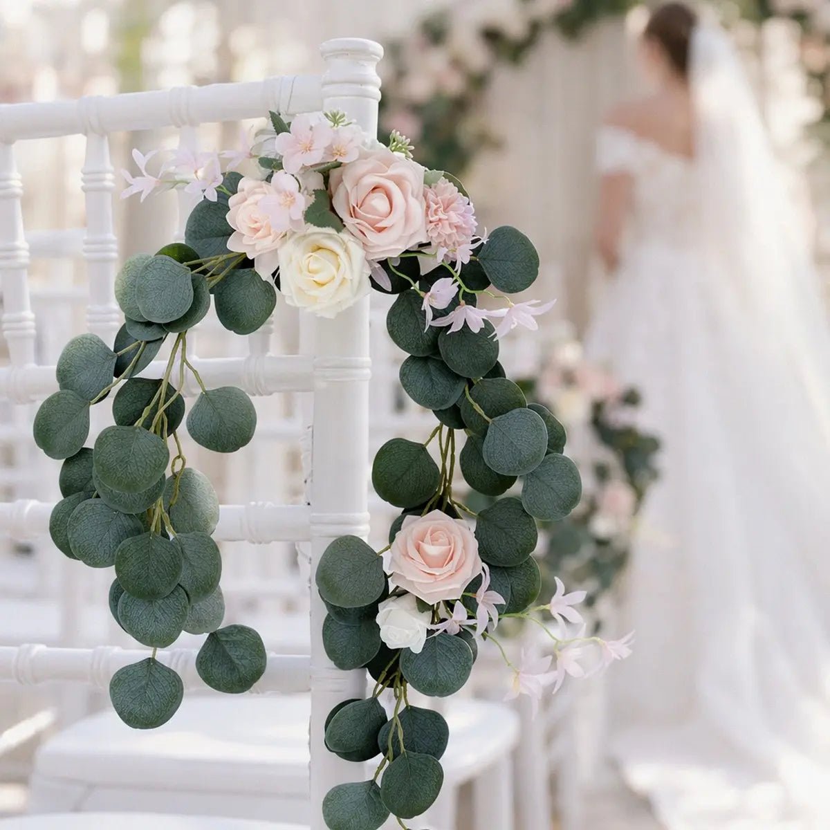 Blush pink rose silver dollar eucalyptus chair swag draped on white Chiavari aisle chair back with bride in soft focus background at indoor wedding ceremony – IPOPU