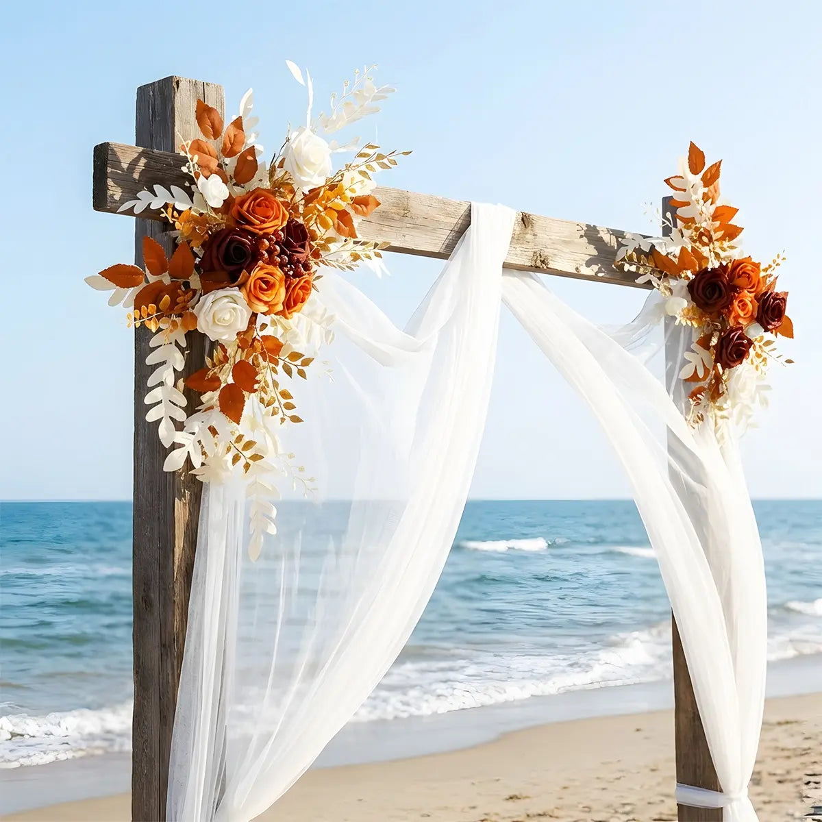 Burnt orange artificial flower swags decorating a beachside wedding arch with white draped fabric
