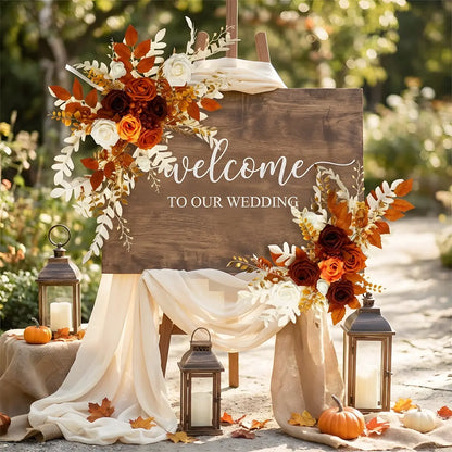 Burnt orange boho artificial flower swag decorating a rustic wedding welcome sign with lanterns and draped fabric
