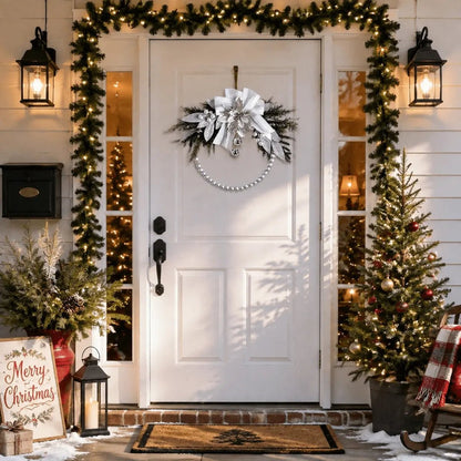 Silver white Christmas swag wreath with oversized ribbon bow and pearl chain hanging on white front door with lit outdoor garland, lanterns, and snow-dusted Christmas trees at night
