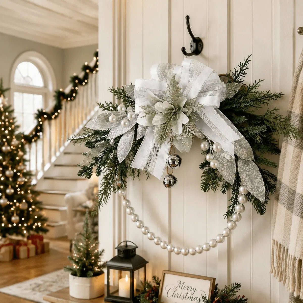 Silver white Christmas swag wreath with ribbon bow, glitter poinsettia, jingle bells, and pearl bead chain hanging on white shiplap wall in festive entryway with staircase garland and lit Christmas tree
