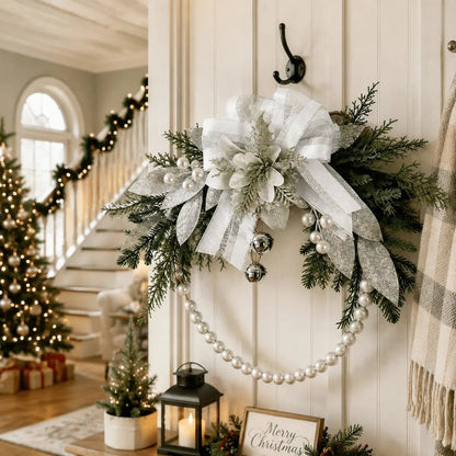 Silver white Christmas swag wreath with ribbon bow, glitter poinsettia, jingle bells, and pearl bead chain hanging on white shiplap wall in festive entryway with staircase garland and lit Christmas tree
