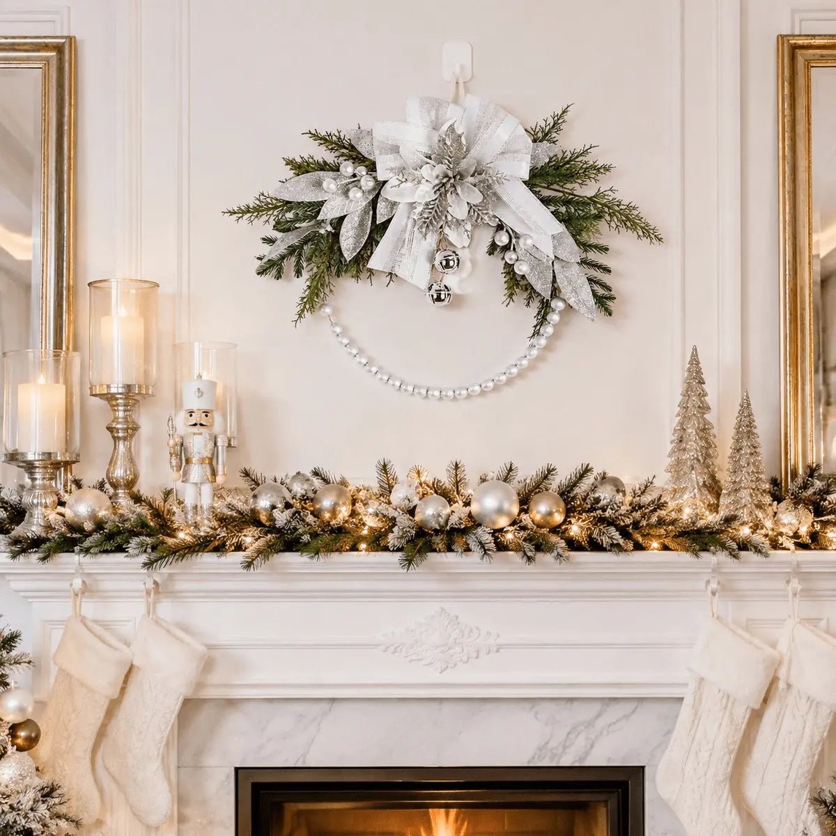 Silver white Christmas swag wreath with glitter poinsettia, ribbon bow, jingle bells, and pearl bead swag displayed above fireplace mantel with white stockings, candles, and nutcracker figurine
