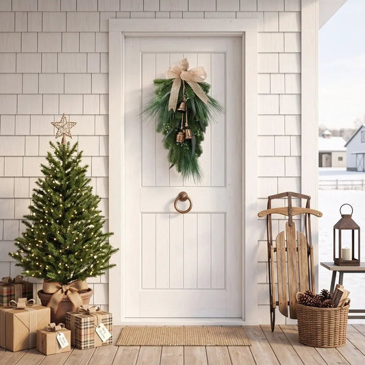 Christmas pine needle teardrop swag with burlap bow and hanging vintage brass bells, displayed on a white shingle farmhouse front door, with a lit mini Christmas tree, kraft paper wrapped gifts and a wooden sled with pine cones on the snowy porch

